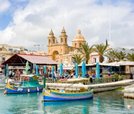 Strona główna Colorful fishing boats and lively market in Marsaxlokk, Malta, with a historic church in the background.